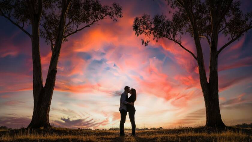 A newly engaged couple sharing a passionate kiss at sunset, silhouetted against the vibrant orange sky over Heathmont engagement photography picturesque locations, with tall eucalyptus trees framing the dramatic scene.