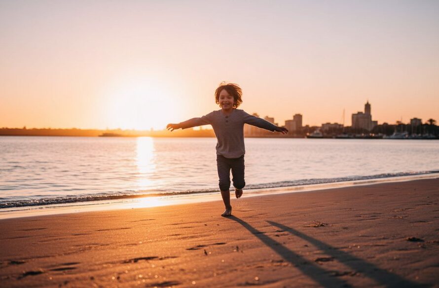 A wide-angle, vibrant photograph capturing joyful child photos East Geelong waterfront, featuring a young girl mid-jump with arms outstretched, silhouetted against a golden sunset over Corio Bay, a blurred waterfront park in the background, conveying pure delight and freedom.
