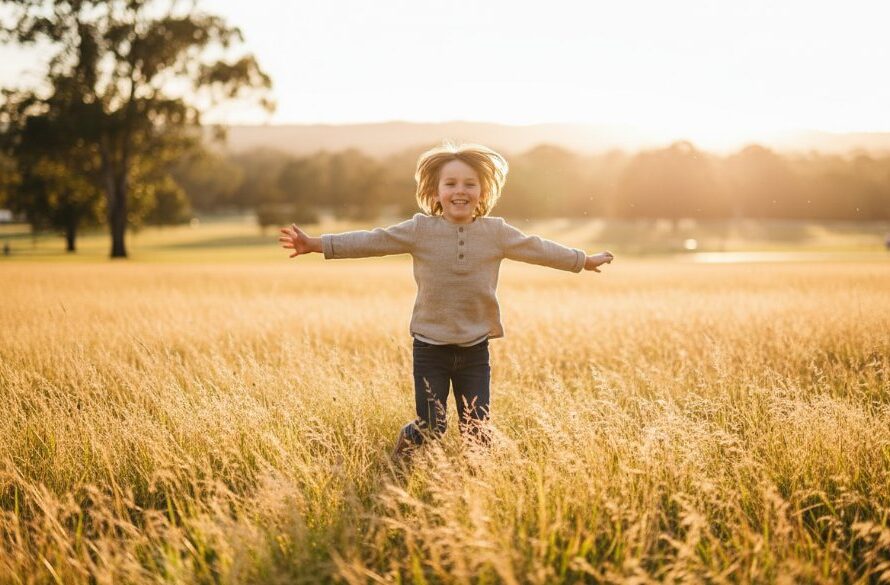 A wide-angle, vibrant, professional photograph capturing the pure joy of a child mid-laugh, running through sun-drenched long grass at a local park in Moe, Victoria. This image epitomises joyful kids photography Moe Victoria outdoor, with warm light highlighting their dynamic movement and genuine happiness.
