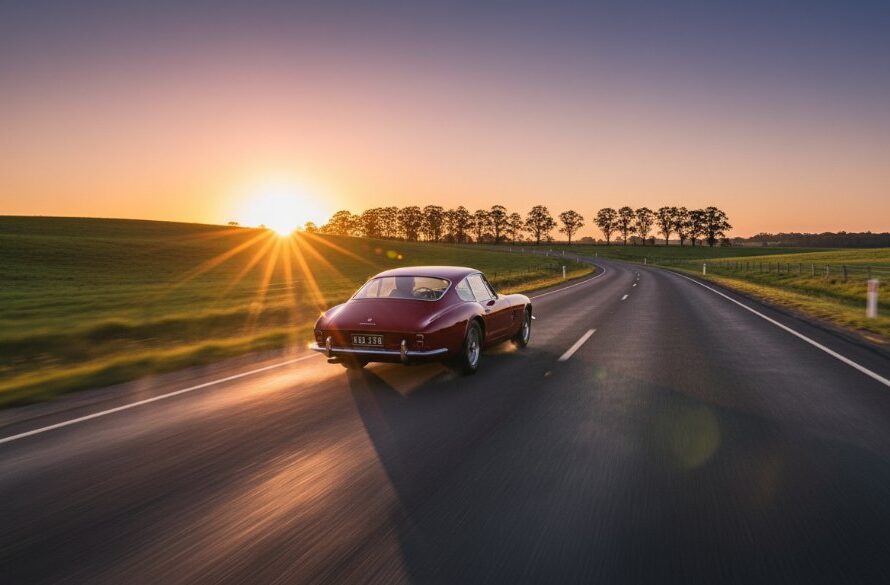 A vintage crimson sports car dramatically posed on a winding, tree-lined road at sunset, capturing the essence of Korumburra classic car photography scenic drives with golden hour light illuminating its sleek lines.