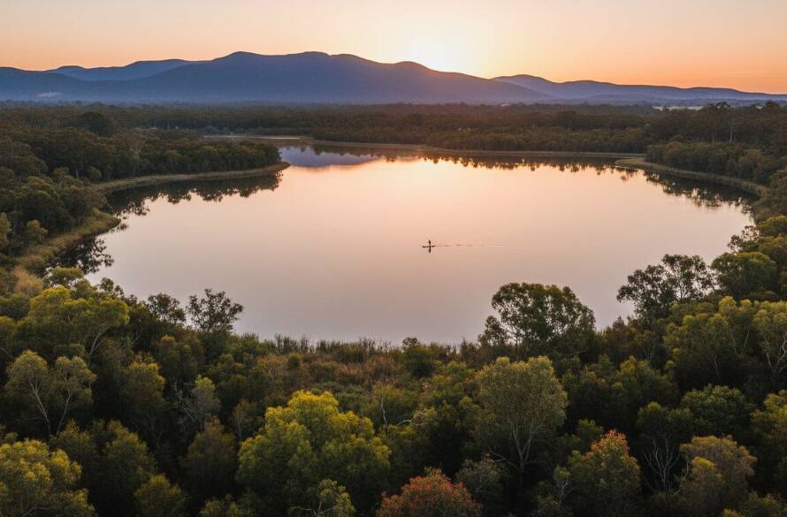 Breathtaking aerial drone shot capturing the serene Lysterfield Lake at sunrise, with a lone stand-up paddleboarder gliding across the calm water, showcasing Lysterfield Lake drone photography services.
