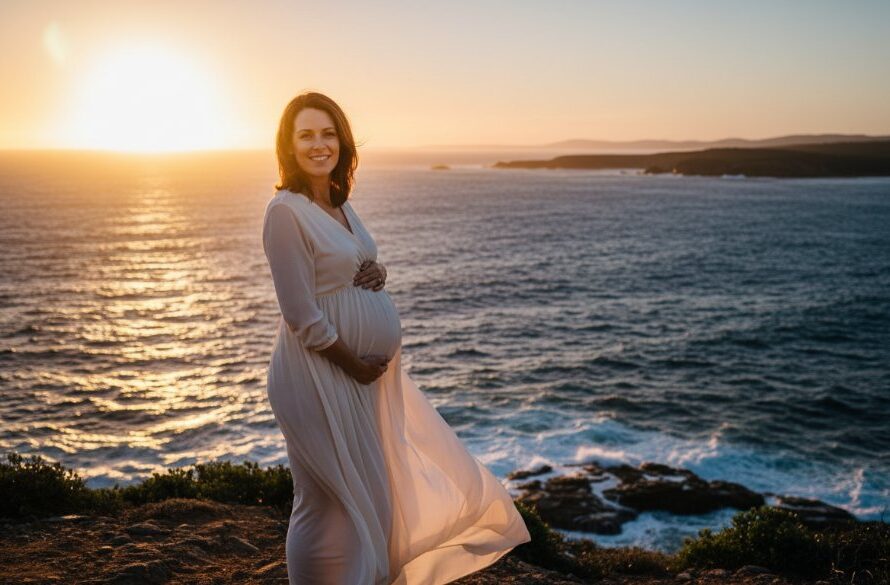 A glowing expectant mother embracing her bump during Mallacoota foreshore maternity photos Victoria, silhouetted against a dramatic sunset at Bastion Point, with waves gently crashing.