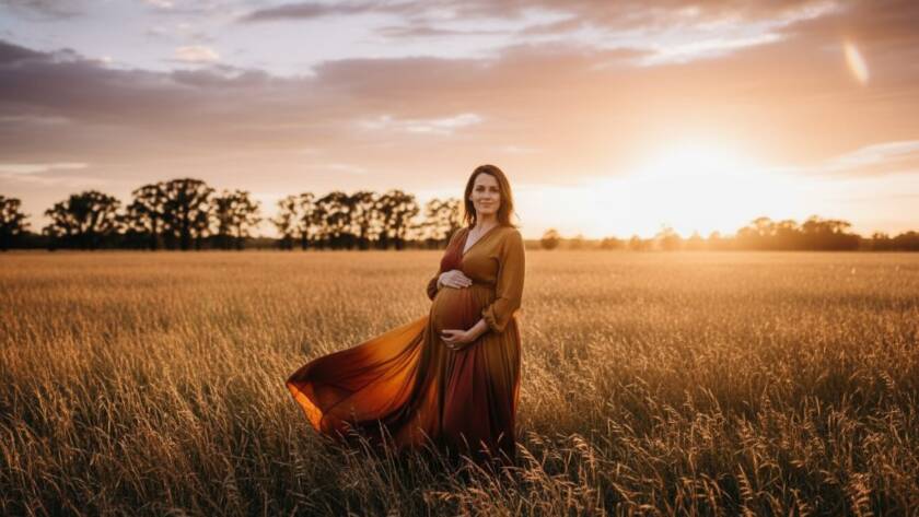 A radiant mother-to-be in an elegant gown, cradling her baby bump, captured during a professional Melton Victoria outdoor maternity photography session at sunset, with dramatic, warm lighting filtering through gum trees.