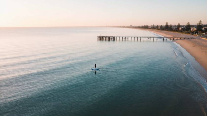An epic aerial shot of Mentone Beach at sunrise, captured by Mentone Coastal Drone Photography, showing golden light illuminating the sandy shore and calm turquoise waters with a lone paddleboarder. The scene exudes serene beauty and professional colour grading.