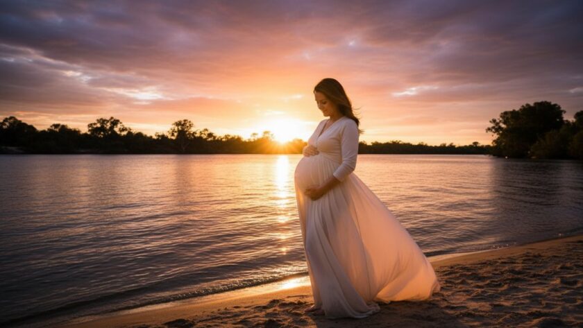 An expectant mother glowing during a Merbein Victoria Maternity Photoshoot Sunset session. She stands by the Murray River, silhouetted against a fiery orange and purple sky, gently cradling her baby bump. The scene is professionally color-graded with dramatic backlighting.