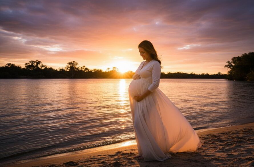 An expectant mother glowing during a Merbein Victoria Maternity Photoshoot Sunset session. She stands by the Murray River, silhouetted against a fiery orange and purple sky, gently cradling her baby bump. The scene is professionally color-graded with dramatic backlighting.