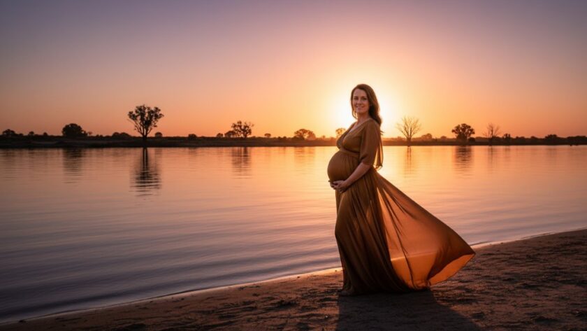 A breathtaking wide shot of a glowing expectant mother at golden hour during a Mildura maternity photography riverside sunset session, standing gracefully by the Murray River, her silhouette framed by the vibrant sky and soft waters, capturing an epic moment of serene anticipation.