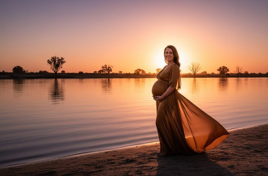 A breathtaking wide shot of a glowing expectant mother at golden hour during a Mildura maternity photography riverside sunset session, standing gracefully by the Murray River, her silhouette framed by the vibrant sky and soft waters, capturing an epic moment of serene anticipation.
