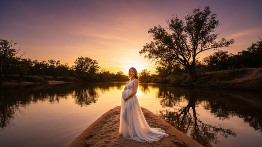 An epic wide-shot Moama maternity photography riverland portraits, featuring a pregnant woman in a flowing gown standing gracefully on a sandbank at sunset, with the golden light reflecting off the Murray River and a majestic eucalypt silhouetted in the background, capturing serene expectant joy.