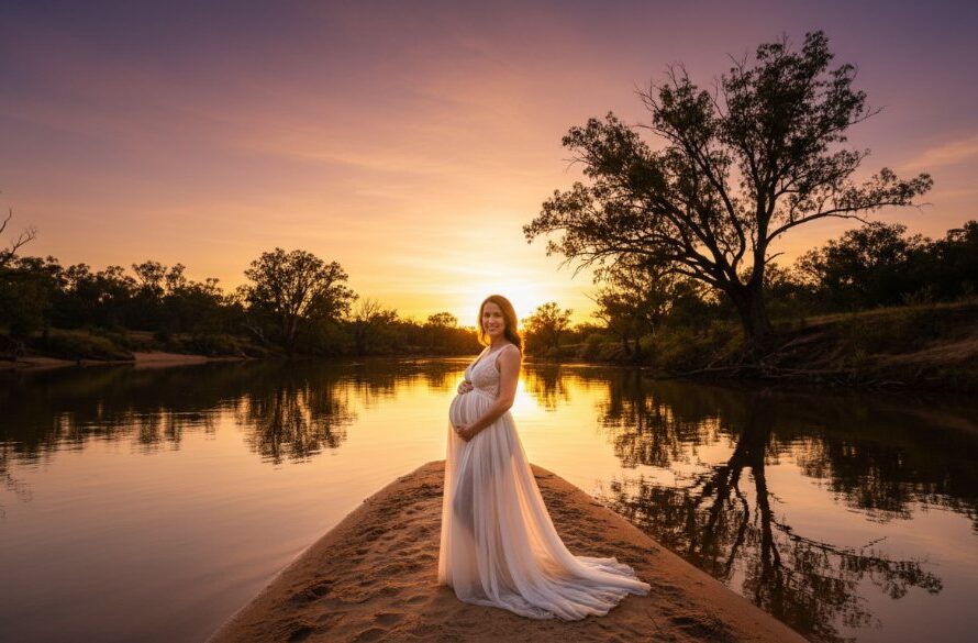 An epic wide-shot Moama maternity photography riverland portraits, featuring a pregnant woman in a flowing gown standing gracefully on a sandbank at sunset, with the golden light reflecting off the Murray River and a majestic eucalypt silhouetted in the background, capturing serene expectant joy.