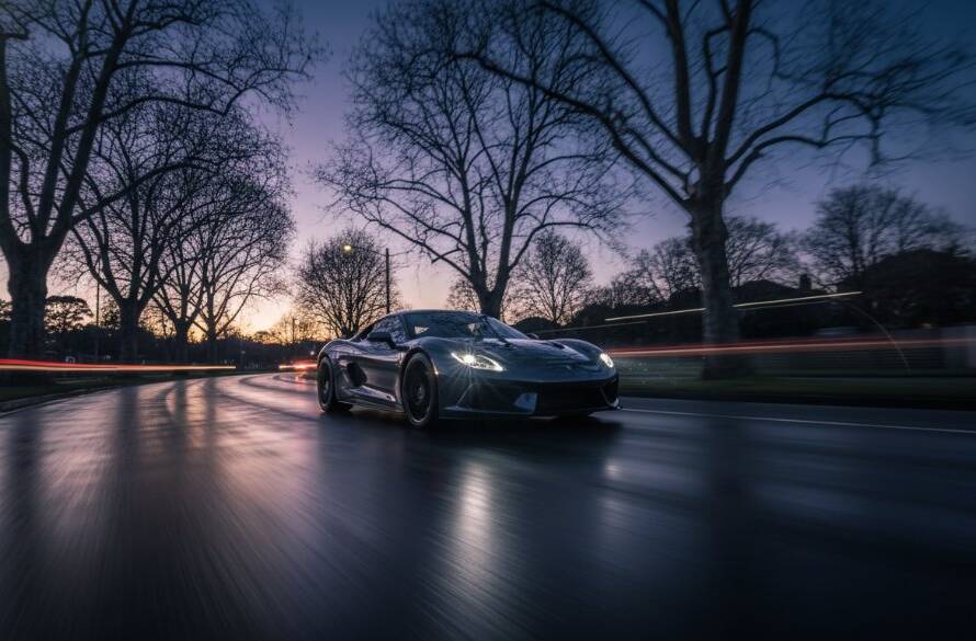 Dramatic low-angle shot of a sleek, dark performance sports car speeding through a scenic tree-lined street in Mont Albert during twilight, showcasing its dynamic lines and the glow of its headlights, epitomizing Mont Albert performance car photography.