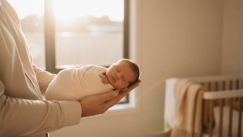 A breathtaking wide shot of a Mordialloc newborn photography natural light session, showcasing a peaceful baby nestled in parents' arms by a sun-drenched window, bathed in soft, golden light, evoking warmth and connection.