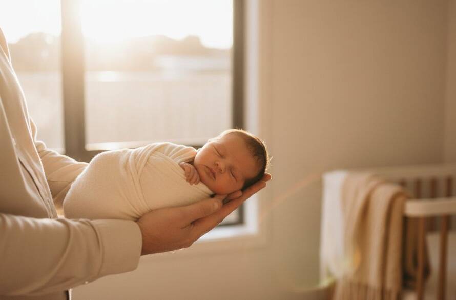 A breathtaking wide shot of a Mordialloc newborn photography natural light session, showcasing a peaceful baby nestled in parents' arms by a sun-drenched window, bathed in soft, golden light, evoking warmth and connection.