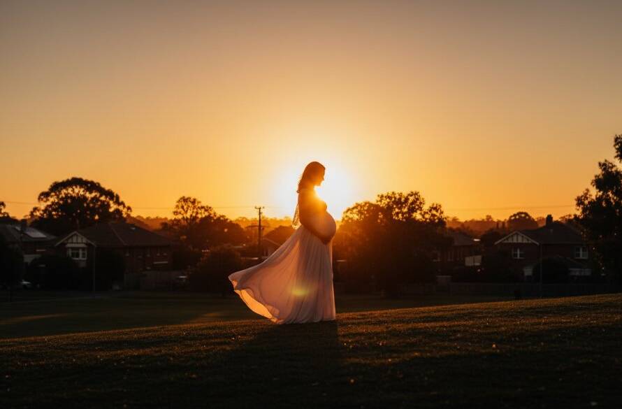 An expectant mother in Murrumbeena, bathed in the soft, golden light of a setting sun, her silhouette creating a dramatic and emotional Murrumbeena maternity photography radiant glow. She stands gracefully by a historic building, gently cradling her baby bump, with a serene expression, professionally colour-graded for a cinematic feel.