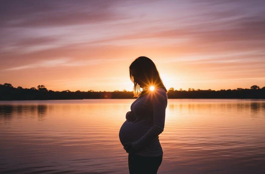 A pregnant woman, looking serene and radiant, stands silhouetted against a golden sunset in a natural setting near Ruffey Lake Park in Doncaster East, Victoria, during her Radiant Doncaster East Maternity Photoshoot Victoria. Her belly is gently cradled, with soft, dramatic backlighting creating a halo effect around her, evoking a sense of powerful calm and anticipation.