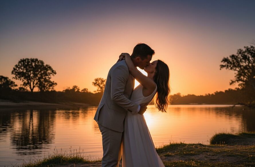 A couple sharing a romantic kiss at sunset by the Murray River, backlit by golden hour glow, with their wedding outfits silhouetted against the water, captured with a cinematic style. This stunning image exemplifies romantic Moama riverside wedding photography.