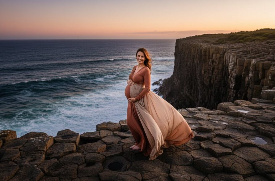 A pregnant woman, in a flowing dress, stands silhouetted against a dramatic sunset over the rugged cliffs of Cape Nelson, embodying a serene Portland Victoria maternity photography experience, with golden light illuminating her profile and the vast ocean stretching behind her.