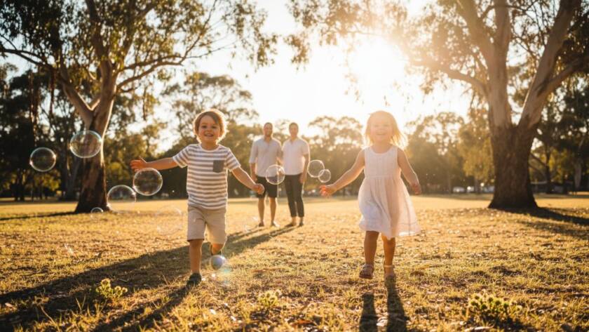 A wide, low-angle shot capturing a joyous St Albans kids photography vibrant outdoor family portrait, with two children (aged 5 and 7) laughing as they chase bubbles in a sun-drenched park at golden hour, their parents smiling softly in the background, creating an epic moment of family connection and playfulness, professionally colour-graded.