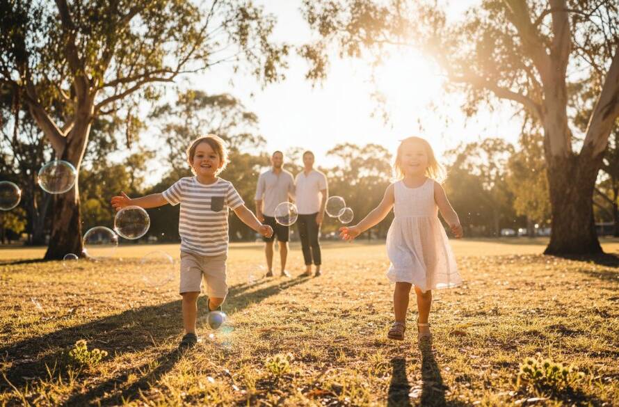 A wide, low-angle shot capturing a joyous St Albans kids photography vibrant outdoor family portrait, with two children (aged 5 and 7) laughing as they chase bubbles in a sun-drenched park at golden hour, their parents smiling softly in the background, creating an epic moment of family connection and playfulness, professionally colour-graded.