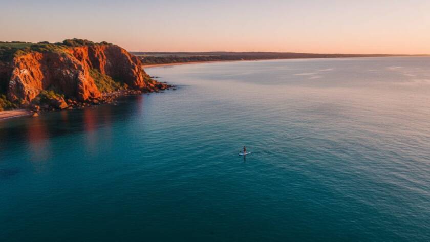 An epic moment shot of stunning Beaumaris coastal drone photography, capturing a lone paddleboarder gliding over crystal-clear turquoise waters near the dramatic red cliffs of Beaumaris Bay, bathed in warm golden hour light, offering a unique aerial view.