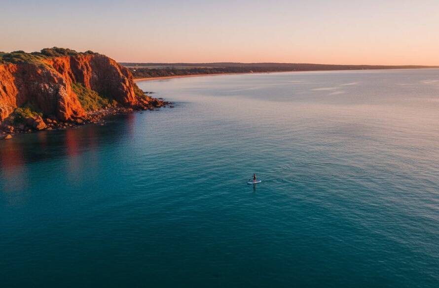 An epic moment shot of stunning Beaumaris coastal drone photography, capturing a lone paddleboarder gliding over crystal-clear turquoise waters near the dramatic red cliffs of Beaumaris Bay, bathed in warm golden hour light, offering a unique aerial view.