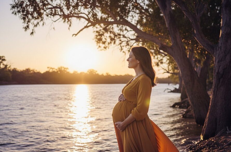 A radiant expectant mother, beautifully silhouetted against a golden hour sunset over the Murray River, during a Swan Hill riverside maternity photoshoot Victoria. Her hands gently cup her baby bump, with the soft, dramatic light creating a cinematic, ethereal glow around her, evoking a sense of peaceful anticipation and natural beauty. Professional colour grading.