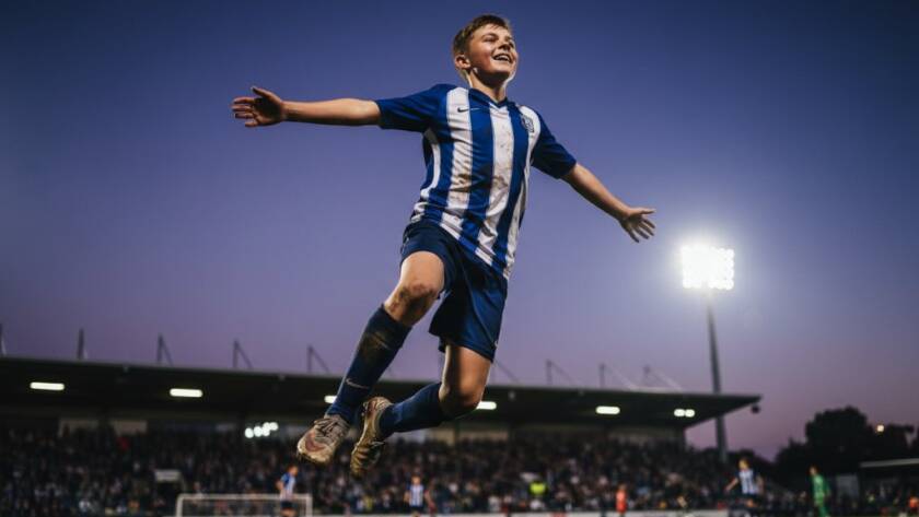 Dynamic wide-angle shot of a young soccer player in mid-kick, mud flying, under dramatic stadium lights during a Templestowe Lower junior sports photography match, capturing an epic goal-scoring moment.