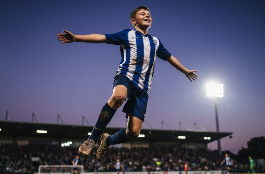 Dynamic wide-angle shot of a young soccer player in mid-kick, mud flying, under dramatic stadium lights during a Templestowe Lower junior sports photography match, capturing an epic goal-scoring moment.