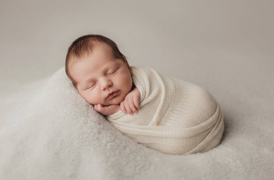 An exquisite, tender newborn photography Burwood portrait capturing a sleeping baby swaddled in soft cream fabric, gently posed in a minimalist studio setting. Soft, dramatic window light illuminates the scene from the side, creating artistic shadows and highlights that emphasize the baby's delicate features, signifying a cherished, peaceful family moment.