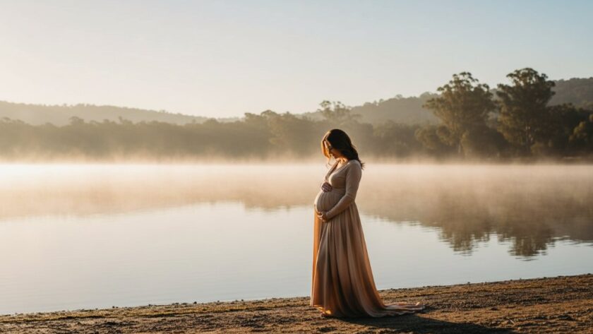A glowing pregnant woman, gracefully posed amidst the soft, ethereal mist of a sunrise over Lake Daylesford, capturing a timeless Hepburn Springs maternity photoshoot experience with dramatic cinematic lighting.