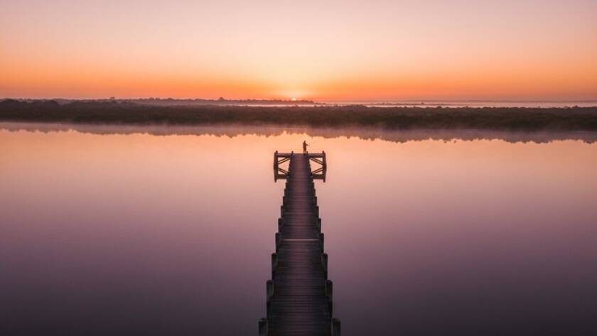 An epic, cinematic wide-angle aerial photograph captured with professional Tooradin Victoria drone photography capturing coastal serenity, showing a lone fisherman casting a line into the calm, glassy waters of Tooradin Jetty at sunrise, with vibrant orange and pink hues reflecting off the water and a dense, misty fog hugging the distant shoreline, evoking a sense of peaceful solitude and dramatic natural beauty, professionally colour graded.