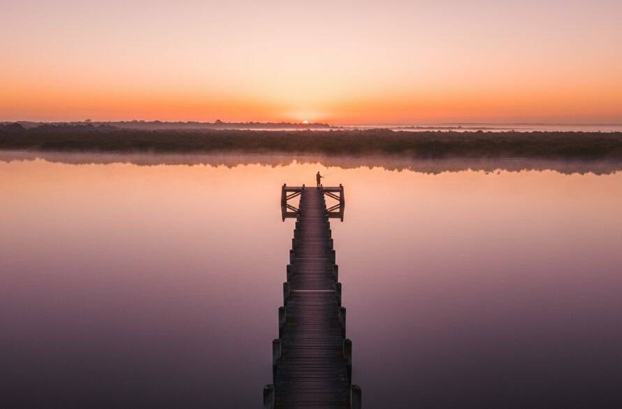 An epic, cinematic wide-angle aerial photograph captured with professional Tooradin Victoria drone photography capturing coastal serenity, showing a lone fisherman casting a line into the calm, glassy waters of Tooradin Jetty at sunrise, with vibrant orange and pink hues reflecting off the water and a dense, misty fog hugging the distant shoreline, evoking a sense of peaceful solitude and dramatic natural beauty, professionally colour graded.