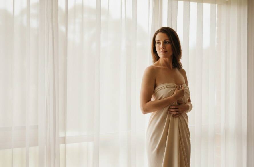 An empowering and beautifully lit portrait of a woman embracing her confidence during a Warrandyte intimate boudoir photography experience, captured in a luxurious, naturally lit room with soft drapes and elegant styling, conveying serenity and strength.