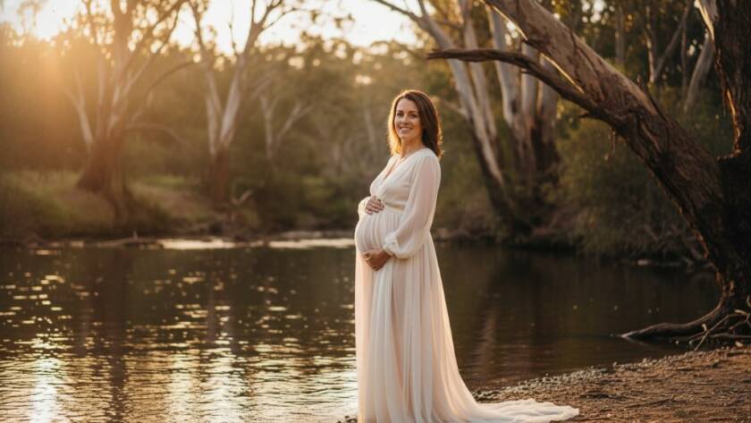 An expectant mother, in a flowing gown, stands gracefully by the tranquil Yarra River in Warrandyte, bathed in the golden light of sunset, showcasing the serene beauty of Warrandyte riverside maternity photography.