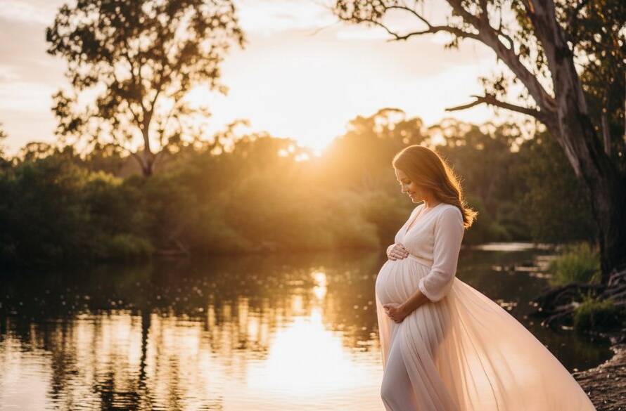 A glowing pregnant woman, in a flowing dress, stands by the Yarra River at sunset in Warrandyte South, her silhouette framed by golden light, epitomising ethereal Warrandyte South Yarra River maternity photography.