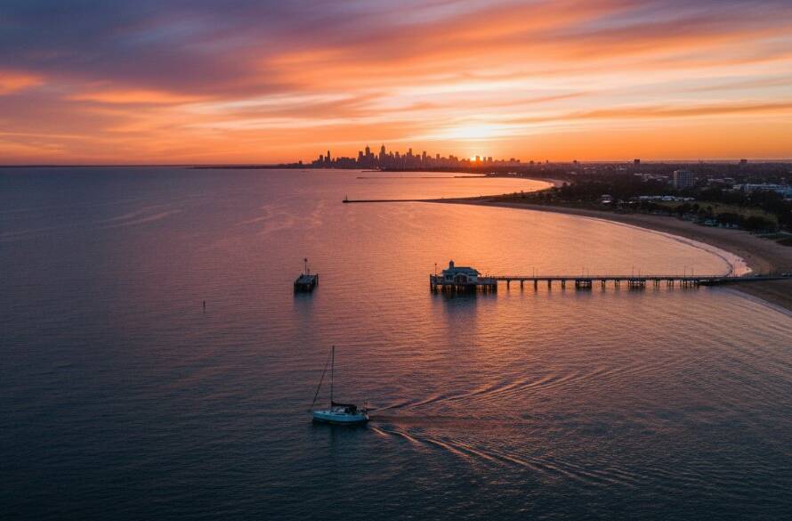 A stunning aerial view showcasing Williamstown drone photography for breathtaking coastal events, capturing a majestic sunset over Williamstown Beach with the Melbourne skyline in the distance, a small sailing boat gracefully cutting through the water, professionally color-graded with dramatic lighting.
