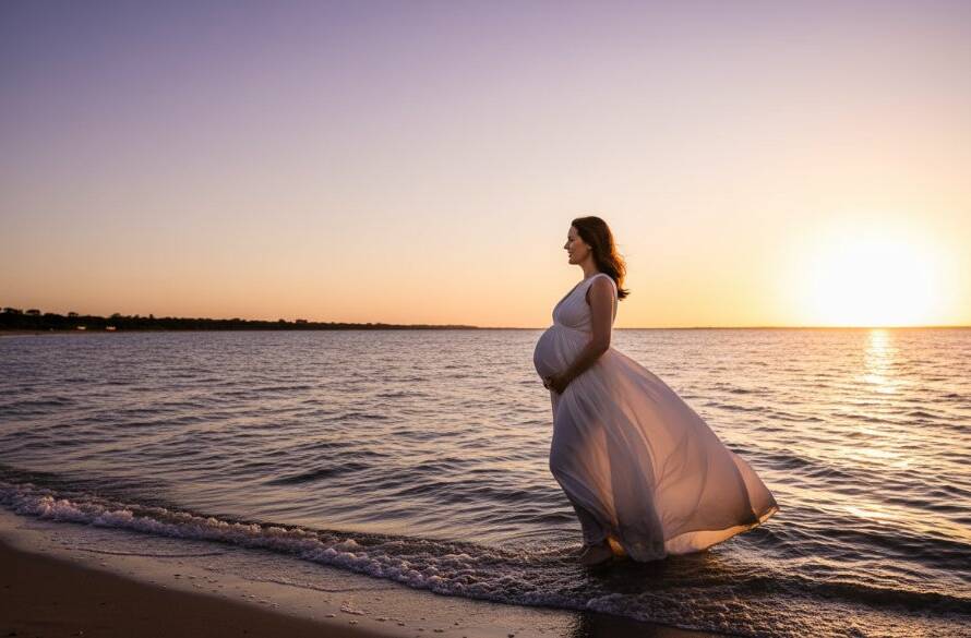 A serene Williamstown North maternity photoshoot capturing a stunning bayside glow at sunset. A radiant pregnant woman in a flowing dress stands silhouetted against the golden hour sky, gentle waves lapping at her feet near Jawbone Marine Sanctuary. Dramatic lighting highlights her silhouette, evoking an epic moment of anticipation and beauty.