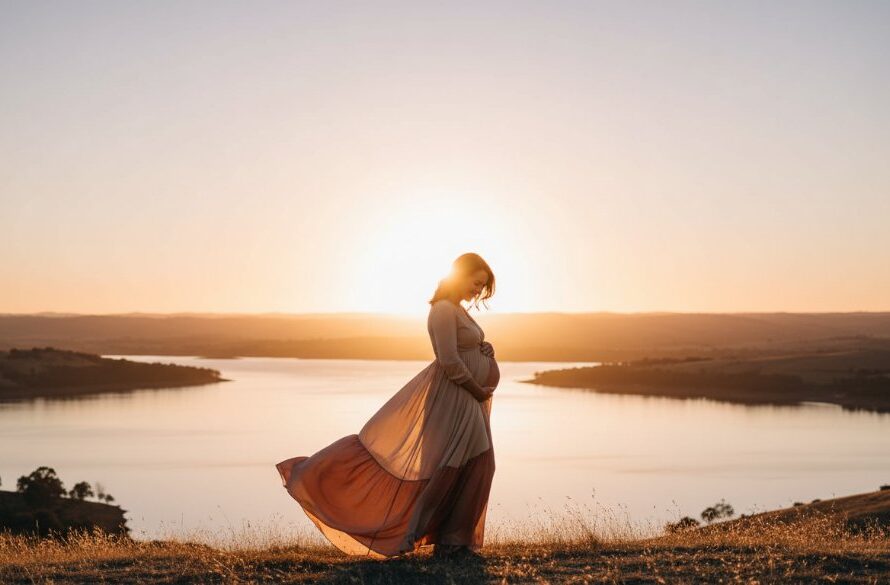 An epic moment captured in Wodonga Victoria elegant maternity photography, featuring a radiant pregnant woman in a flowing gown, silhouetted against a dramatic sunset over the Hume Weir, her hands gently cradling her baby bump.