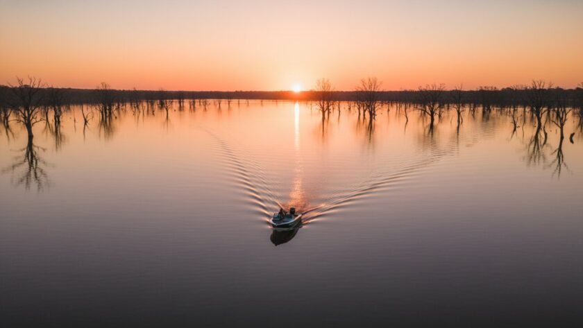 An epic drone shot capturing the serene expanse of Lake Mulwala at sunrise, with a lone boat gliding across the mirror-like surface, showcasing Yarrawonga Drone Photography: Lake Mulwala's Aerial Splendour and the vibrant hues of the dawn sky reflected in the water, framed by distant gum trees.