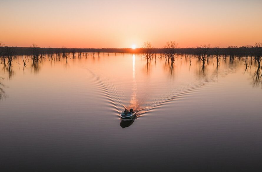 An epic drone shot capturing the serene expanse of Lake Mulwala at sunrise, with a lone boat gliding across the mirror-like surface, showcasing Yarrawonga Drone Photography: Lake Mulwala's Aerial Splendour and the vibrant hues of the dawn sky reflected in the water, framed by distant gum trees.