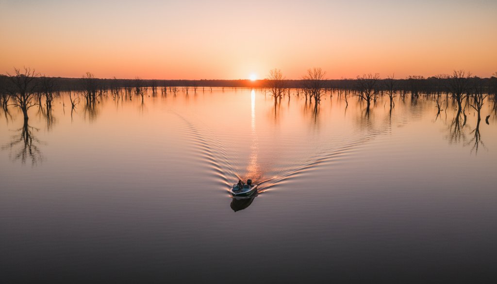 An epic drone shot capturing the serene expanse of Lake Mulwala at sunrise, with a lone boat gliding across the mirror-like surface, showcasing Yarrawonga Drone Photography: Lake Mulwala's Aerial Splendour and the vibrant hues of the dawn sky reflected in the water, framed by distant gum trees.